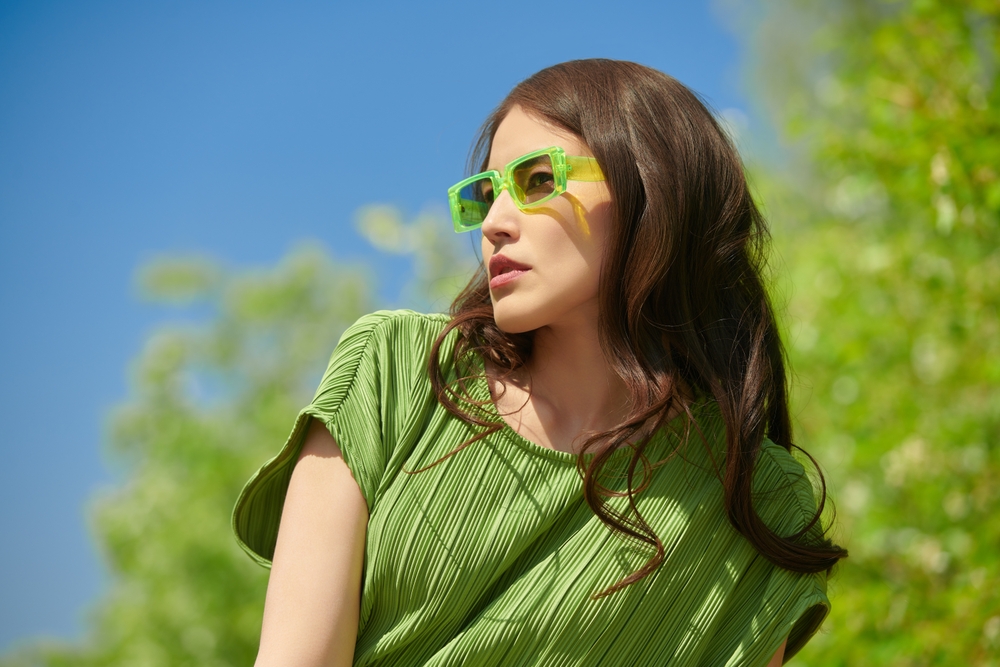 Stylish woman in green sunglasses and dress outdoors on a sunny day – Sunny Vision Corp Fashionable woman wearing bright green sunglasses and a pleated green dress, standing against a clear blue sky and leafy background, looking confidently to the side – Sunny Vision Corp