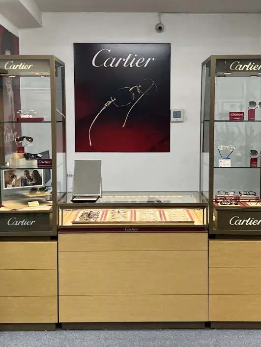 Cartier eyewear display inside optical boutique – Sunny Vision Corp Showcase of luxury Cartier eyeglasses in a retail optical display, with illuminated glass cases on either side and a large Cartier poster featuring elegant rimless frames on the back wall – Sunny Vision Corp