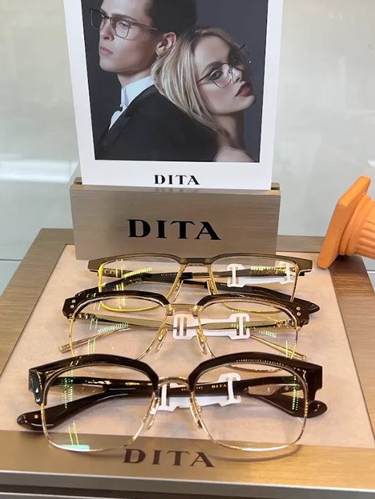 Display of DITA eyeglasses with promotional photo – Sunny Vision Corp Three pairs of stylish DITA eyeglasses with gold and dark frames neatly arranged on a display tray, positioned in front of a branded promotional image featuring a man and woman wearing the glasses – Sunny Vision Corp